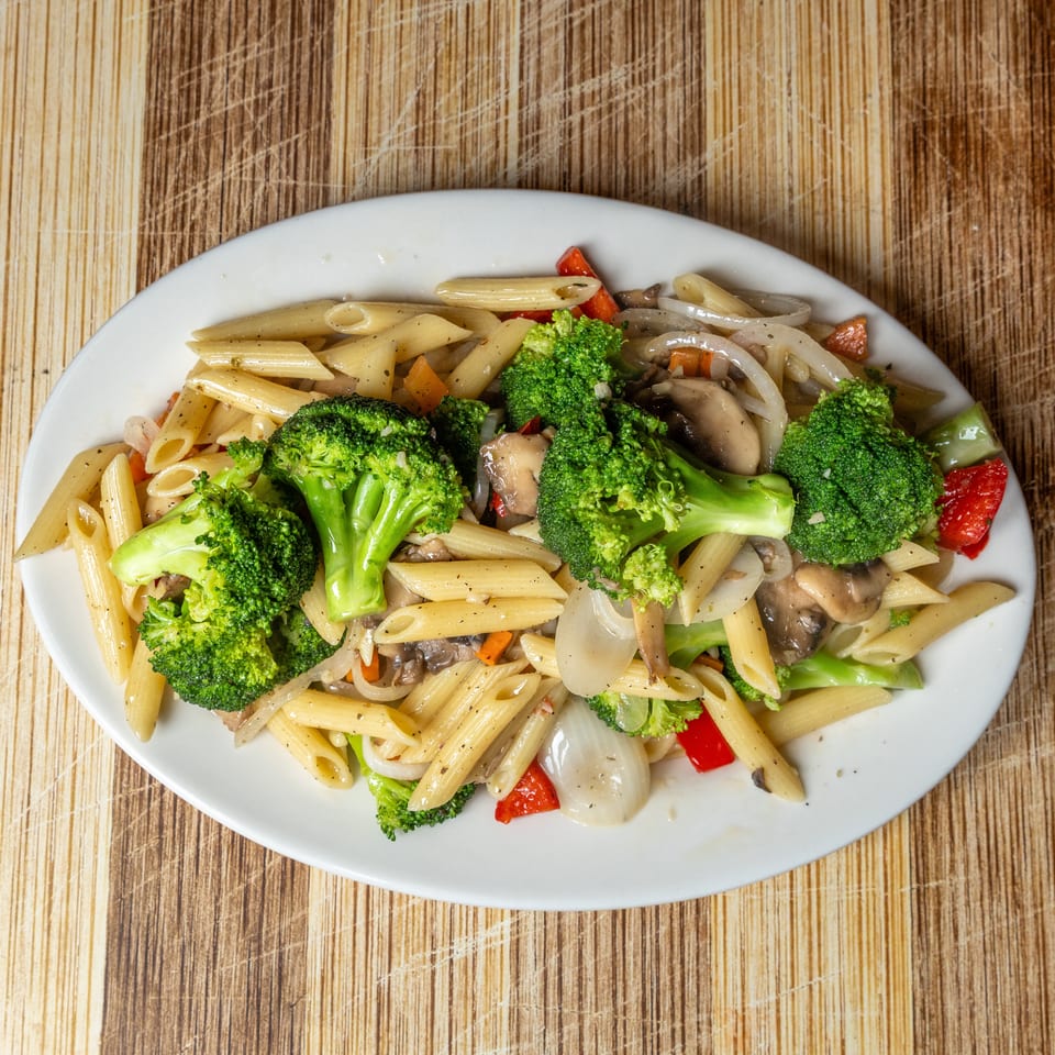 Penne pasta with broccoli, mushrooms, onions, and red bell peppers on a white oval plate.