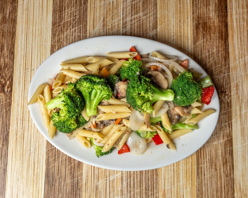 Penne pasta with broccoli, mushrooms, onions, and red peppers on a white oval plate.