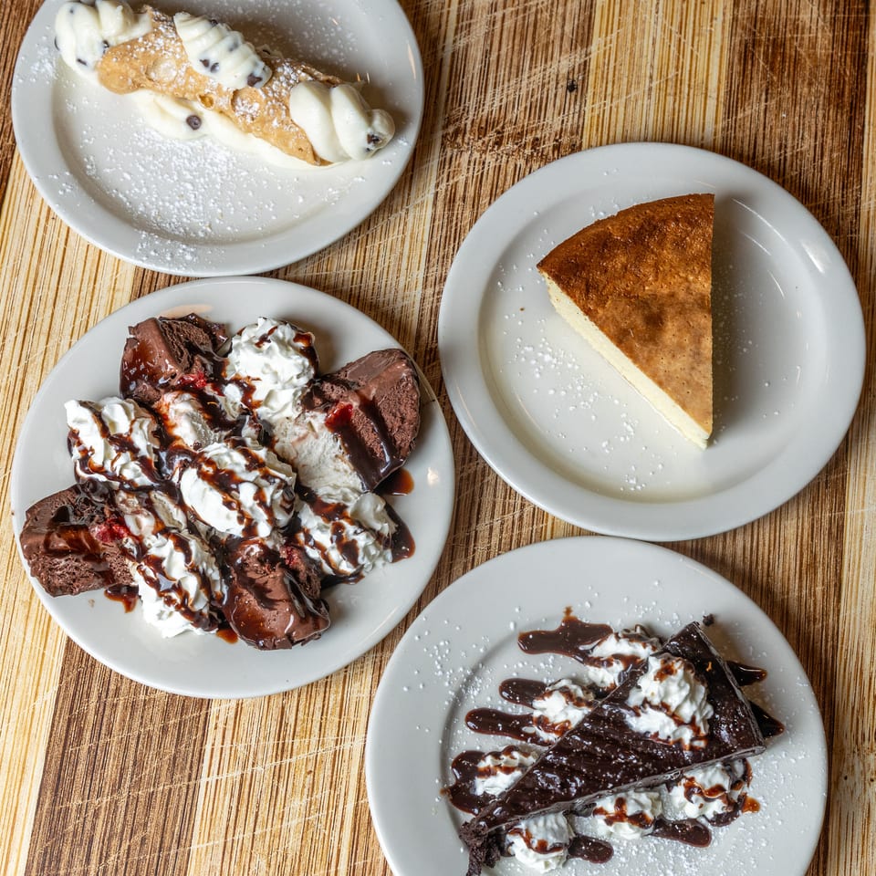 Four dessert plates on a wooden table, including cannoli, cheesecake, and chocolate cake.
